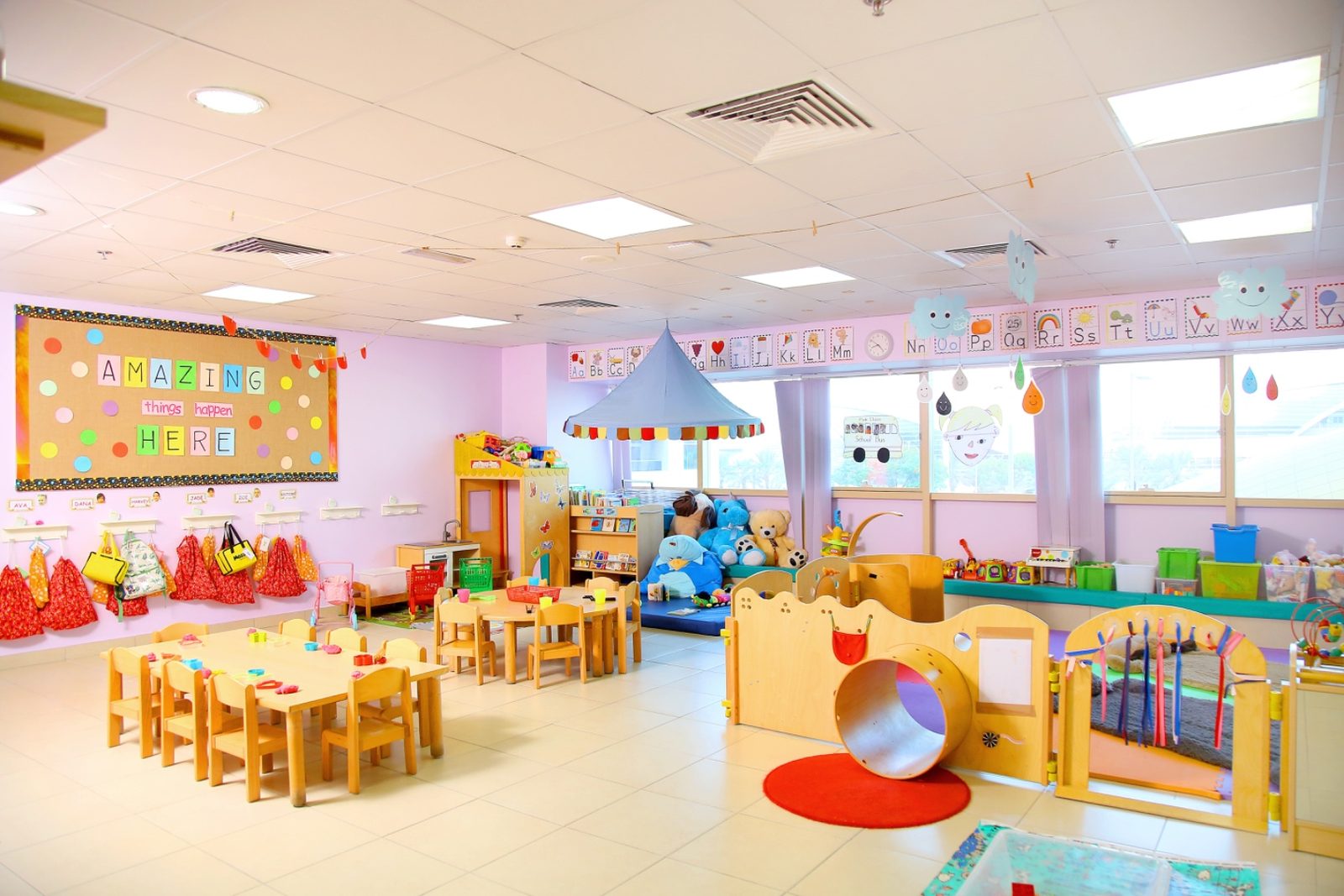 A bright Mosaic Nursery classroom with low colourful shelving, child-height tables and chairs, a small indoor slide and a wall of windows