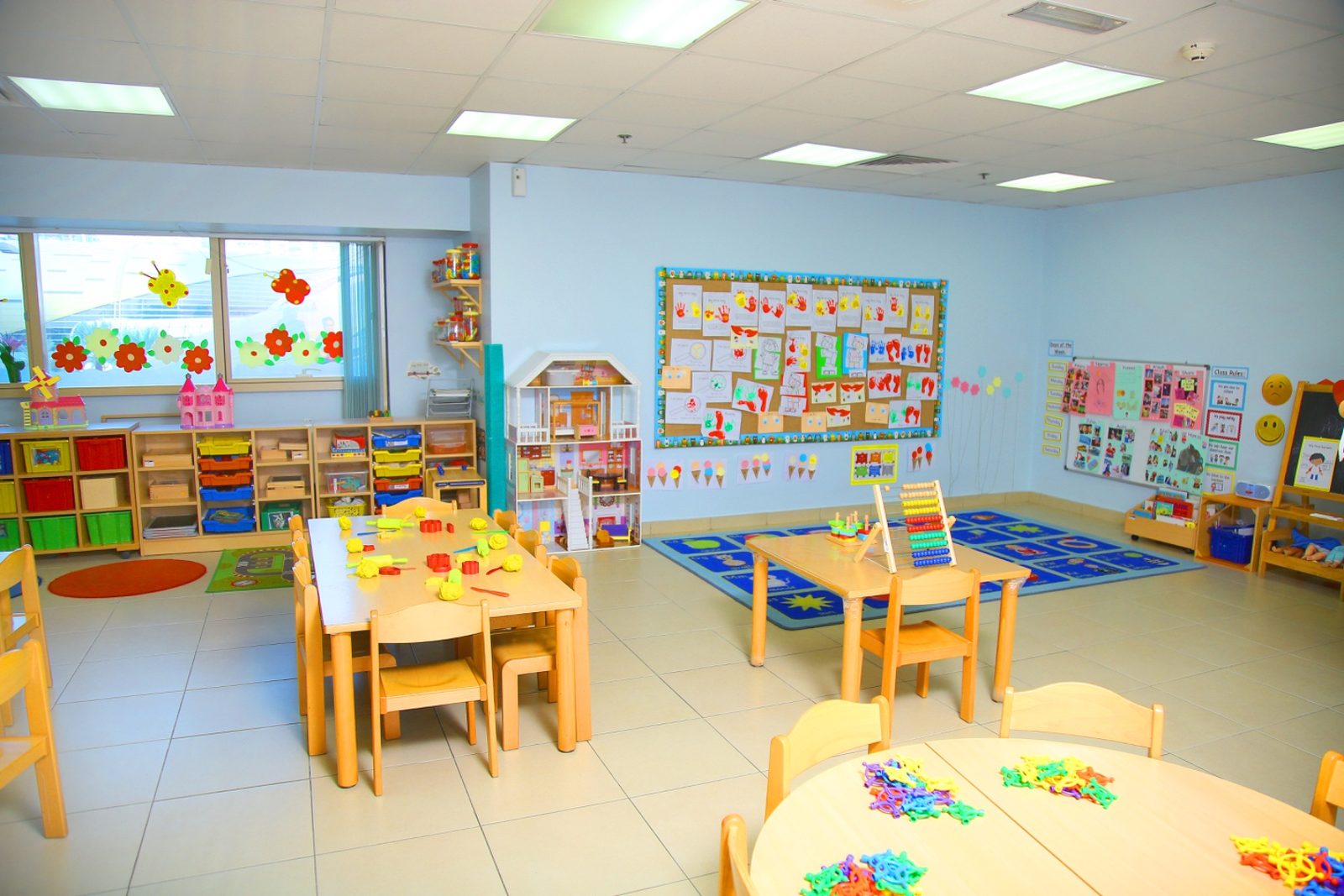 The Pre-KG room at Mosaic, with blue and yellow child-height tables, low shelving, display boards and a wall of windows