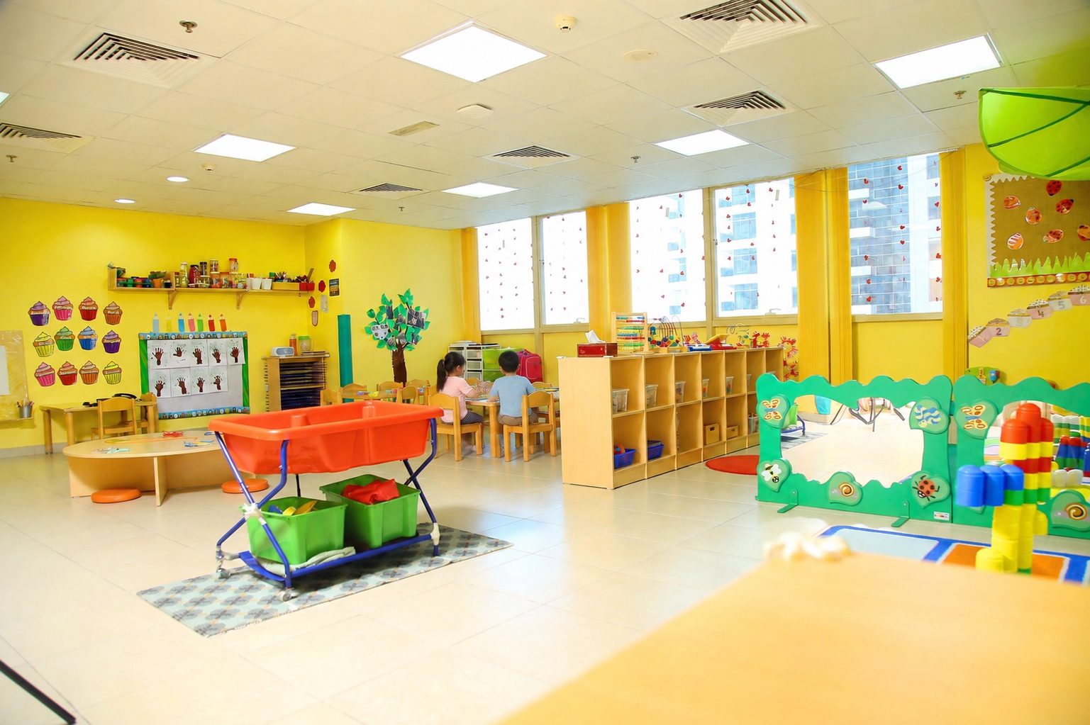 The Toddlers room at Mosaic, with a water tray on a trolley, low shelving of labelled trays, a soft play divider and windows onto the JLT towers